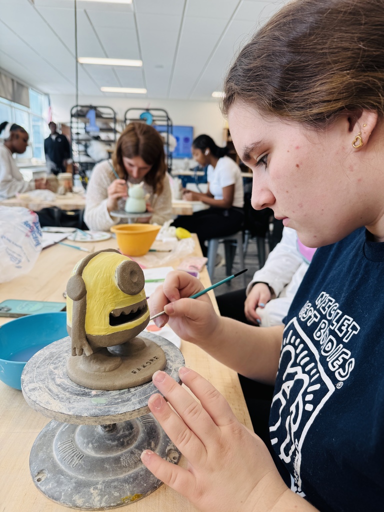 A woman in a blue shirt works on a clay sculpture of a yellow character in a classroom.