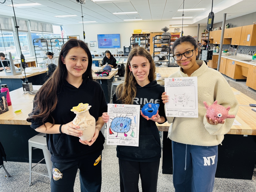 Three girls in a classroom hold items shaped like a virus and a pig. One holds a bag, another a small pig, and the last a paper.