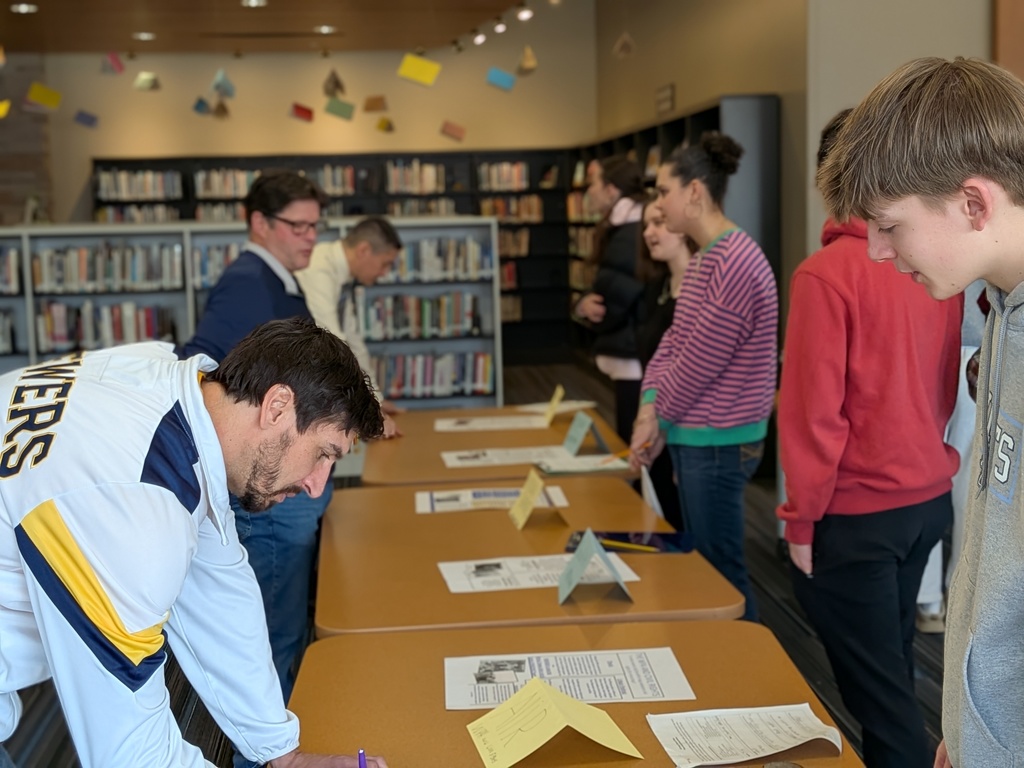 People gathered around tables in a library, with one man signing a paper. Bookshelves in the background.