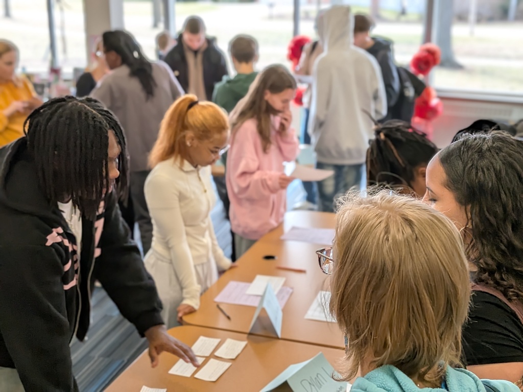 A group of people stand around a table with papers, discussing and pointing. Some wear jackets and glasses.
