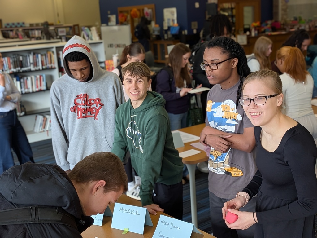 Five people are standing around a table with various items on it in a library.