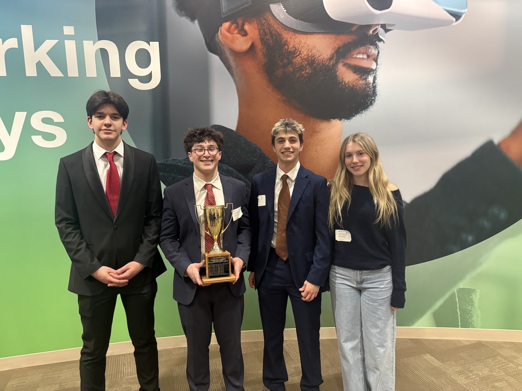 Four people in suits stand in front of a green banner. They are holding a trophy. A person with a beard and virtual reality goggles is on the banner.