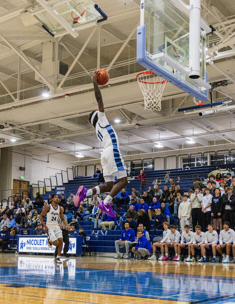 Player in midair, holding a basketball, jumping over another player, aiming for the hoop in an indoor court.