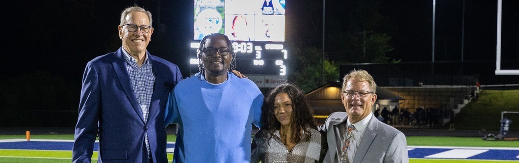 Four people on a football field, smiling for a photo. Background includes a stadium with lights and a scoreboard.
