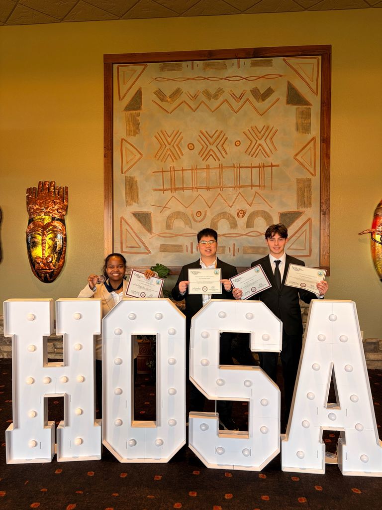Three people stand in front of a lighted HOSA sign. They hold certificates. Behind them is a wall with a large design.