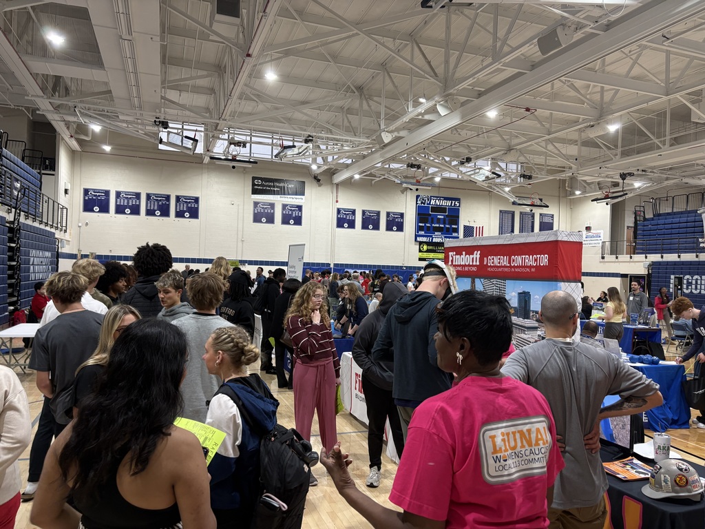Several people gather in a gym for an event with multiple tables and a speaker on the wall.