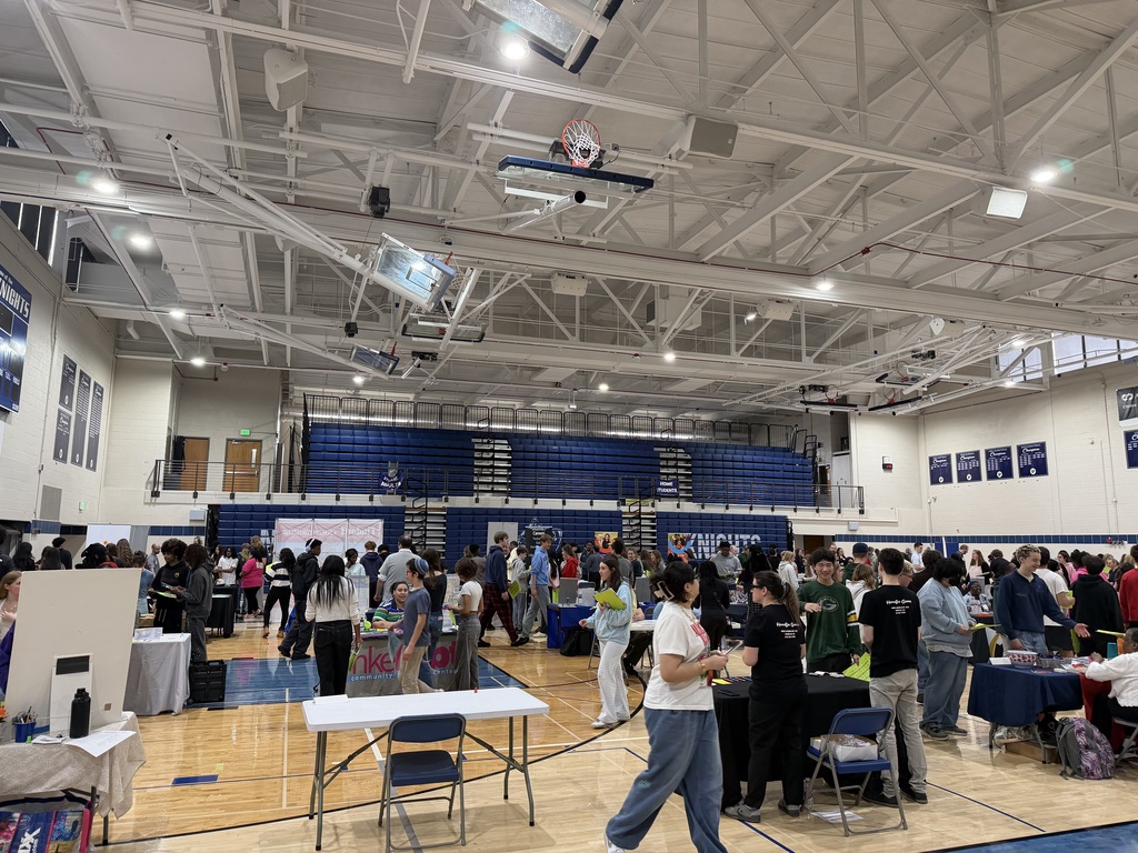 Many people gather in a large indoor space. Tables and chairs are set up around the perimeter. A basketball court is visible in the center.