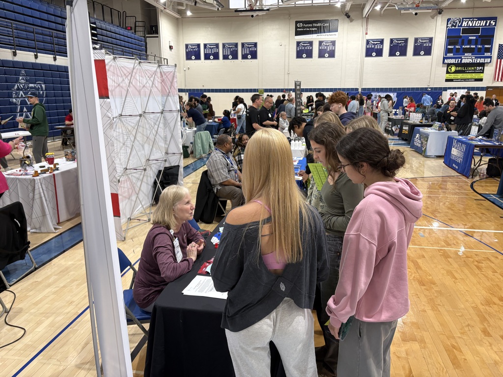 A room with wooden floors, several tables with people standing and sitting around them, likely in an event.