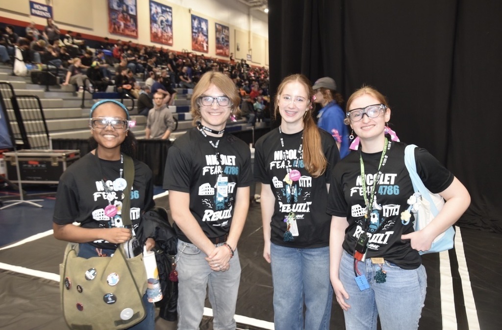 Four individuals wearing black t-shirts and glasses stand together in a room with an audience seated in the background.
