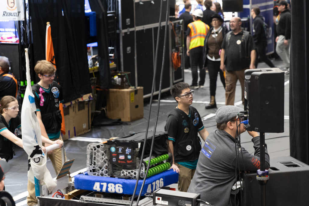 People in a competition area, some with robots, others watching. An orange flag is visible, and a speaker is on a stand.