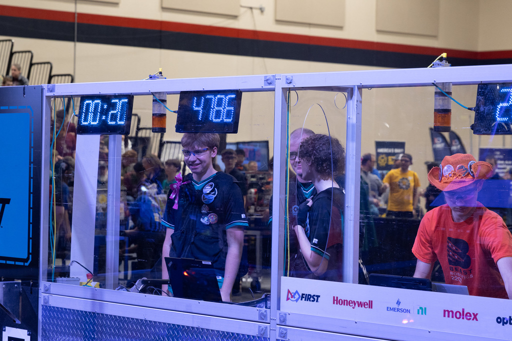 People gathered in a room. Two people wear black shirts, one with a yellow badge. One wears orange.