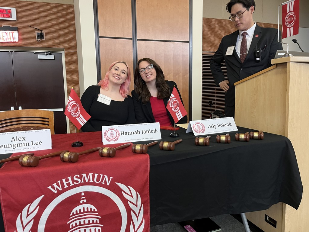 Two people at a table with name placards and flags. A podium is behind them. The table has a WHSMUN logo.