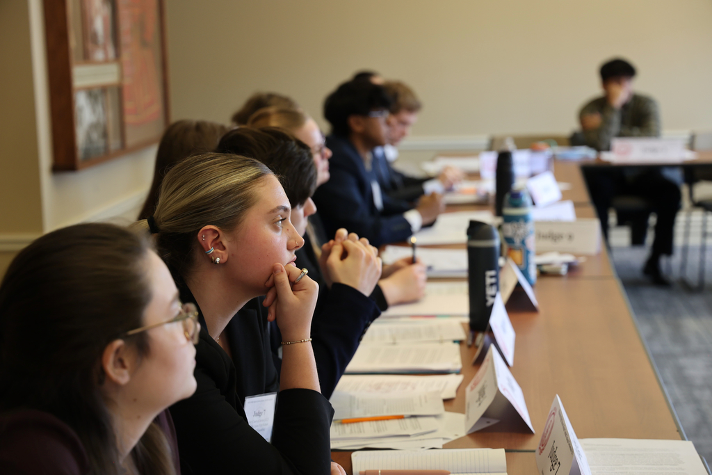 A group of people sits at a long table with papers, pens, and name cards. Some wear glasses.