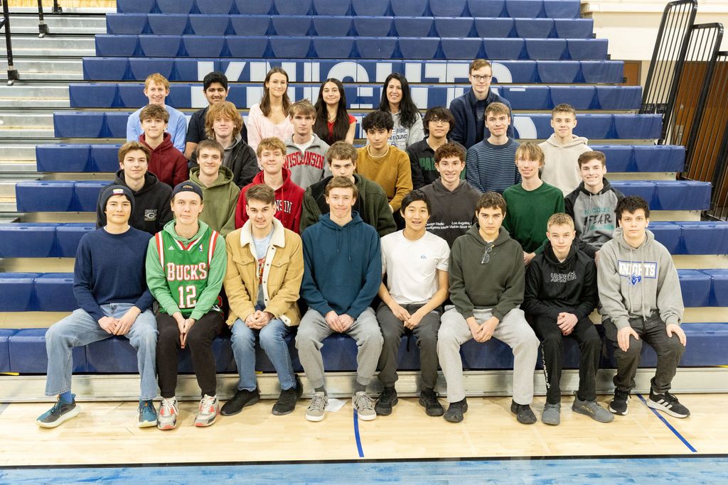 A group of students in a sports uniform sitting in rows on blue bleachers with "Knights" on the wall.