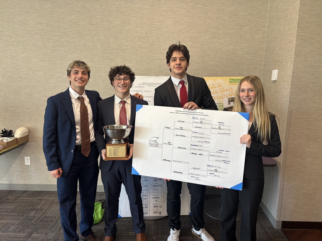 Four individuals stand in a room holding a trophy and a large white paper. One holds the paper while the other holds the trophy.