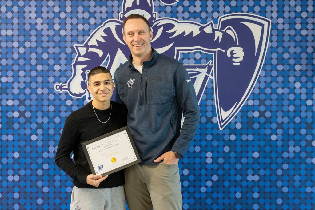Two men stand before a blue background. One holds a framed certificate, and the other has his hands in his pockets.