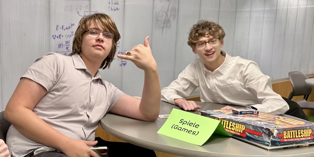 Two students, both wearing glasses, sit at a table. One points up. A board game box labeled "Battleship" sits in front of them.