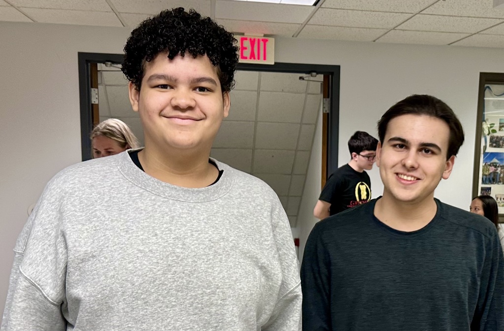 Two young men standing in a hallway, smiling. One wears a gray sweatshirt, the other a black one.