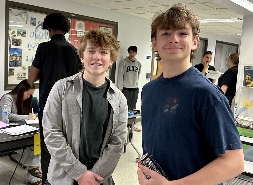 Two boys stand together in a classroom with other students around them. One holds a book, and the other has his hands in his pockets.