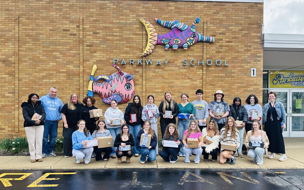 Group of people outside a school with "Parkway School" sign, holding boxes, smiling, and posing for a photo.