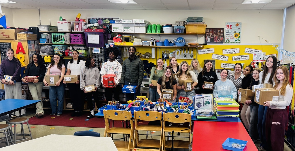 A group of adults and children gather around a table filled with supplies, holding boxes. Shelves in the background are filled with items.
