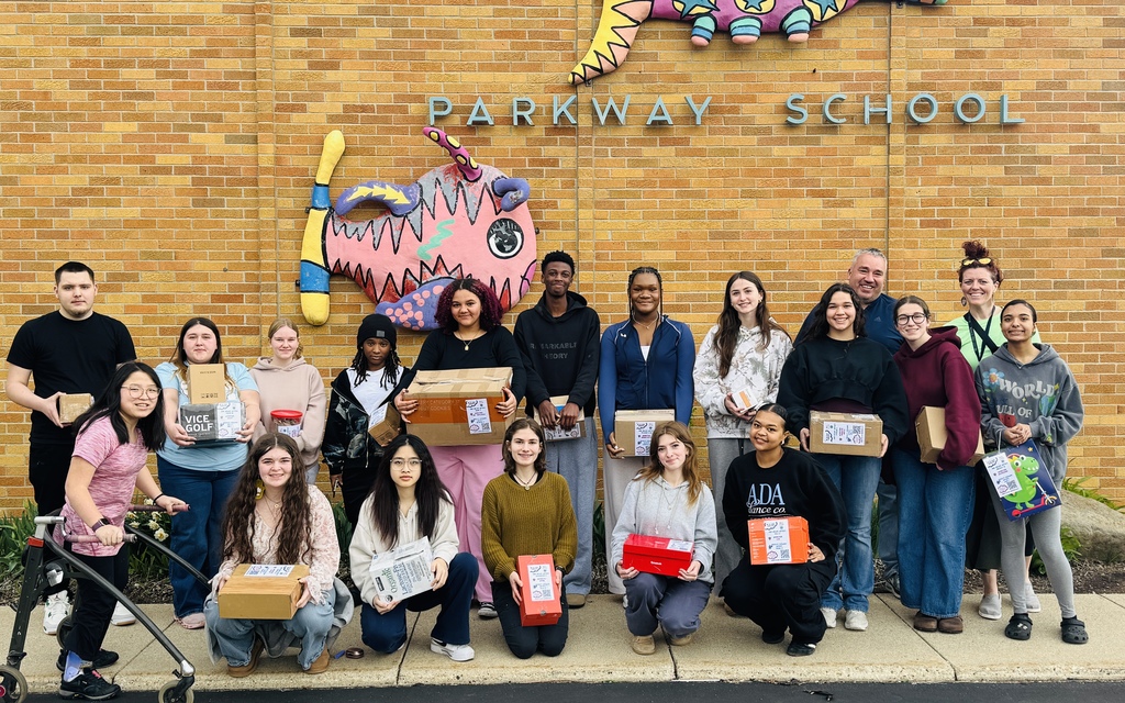 A group of students and staff poses for a photo holding boxes in front of Parkway School's brick building.