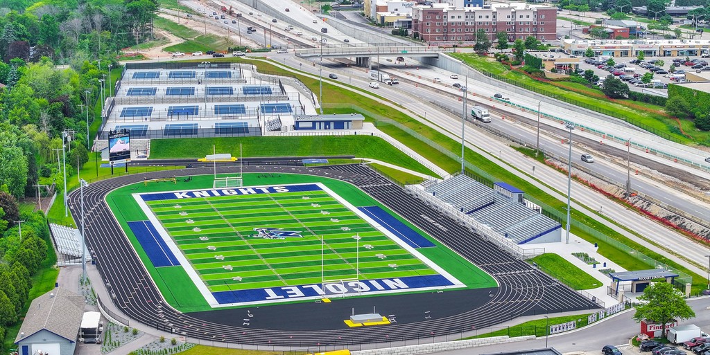 Aerial view of a large football stadium with a blue field, green grass, and parking lot. Several buildings in background.