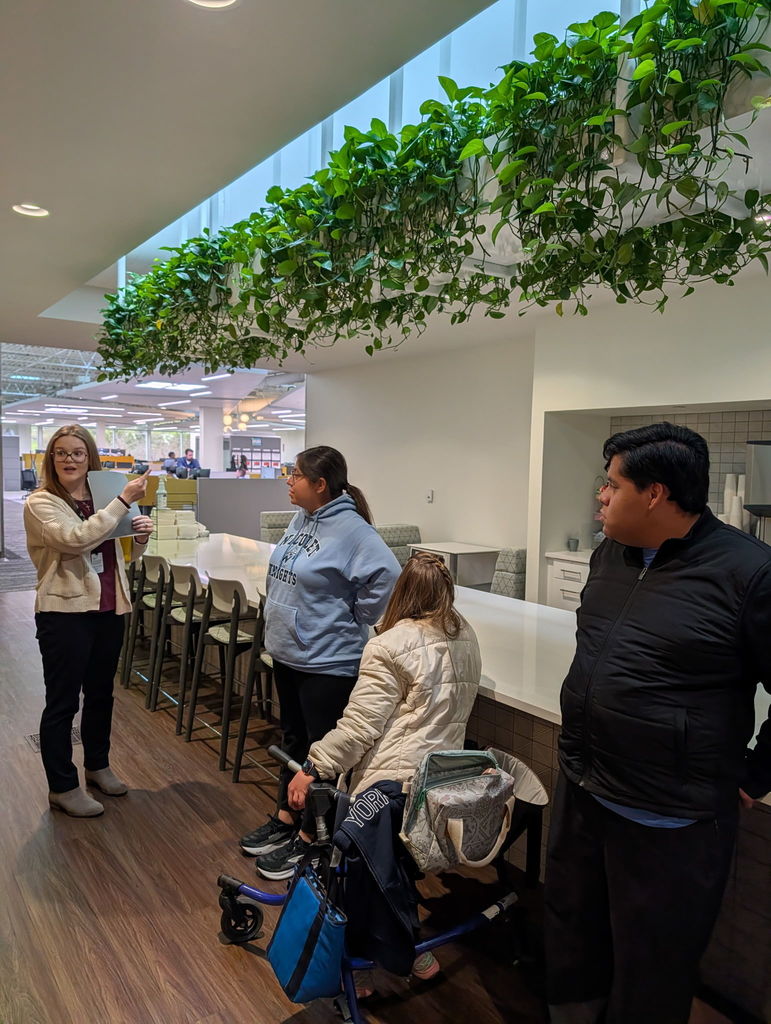 Four individuals stand indoors. One person holds a phone. A person in a wheelchair is near them. Green plants hang from the ceiling.