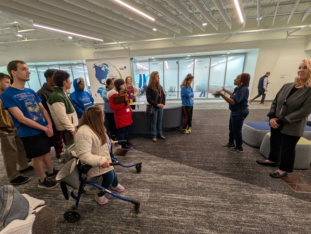 A group of people in a large room with a glass wall. Some are standing, others are sitting, and one person is in a wheelchair.