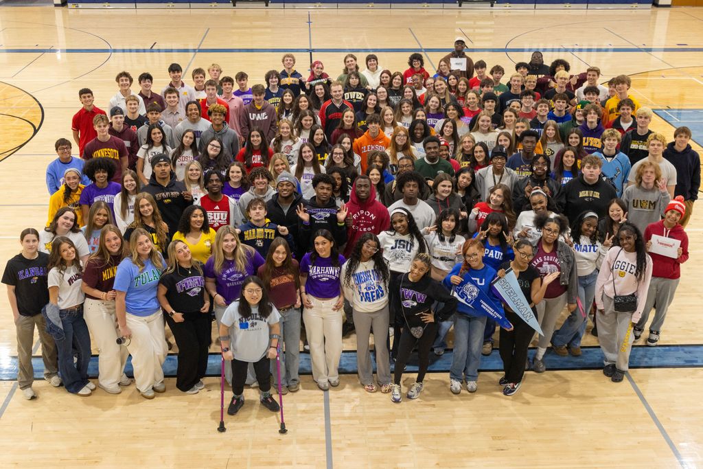 A group of students on a basketball court wearing different colored shirts, standing and sitting, smiling for a photo.