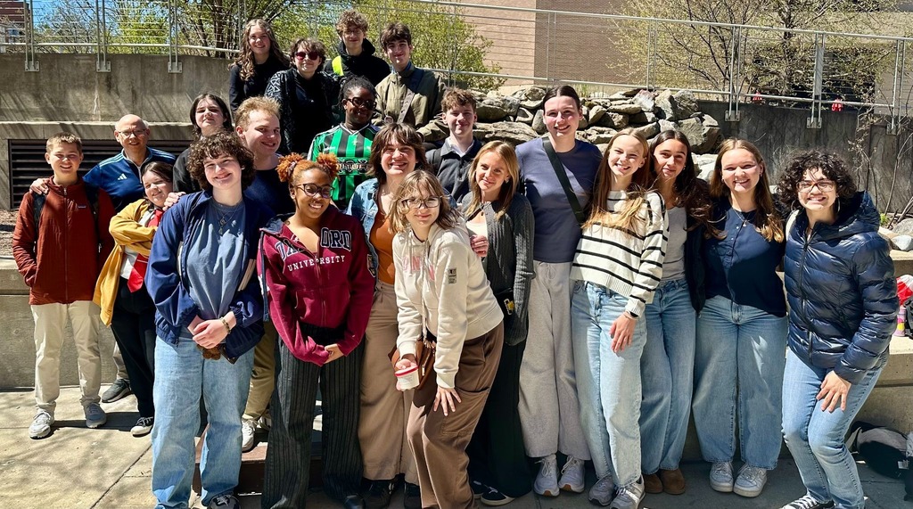 A group of people stands in front of a fountain. Some wear jackets and sneakers, with one woman in a red coat.