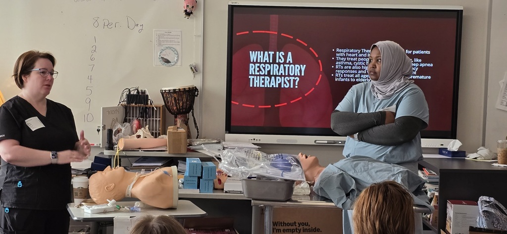 A woman stands in front of a screen with a presentation about respiratory therapists. Another woman in a blue uniform is seated in front of her.