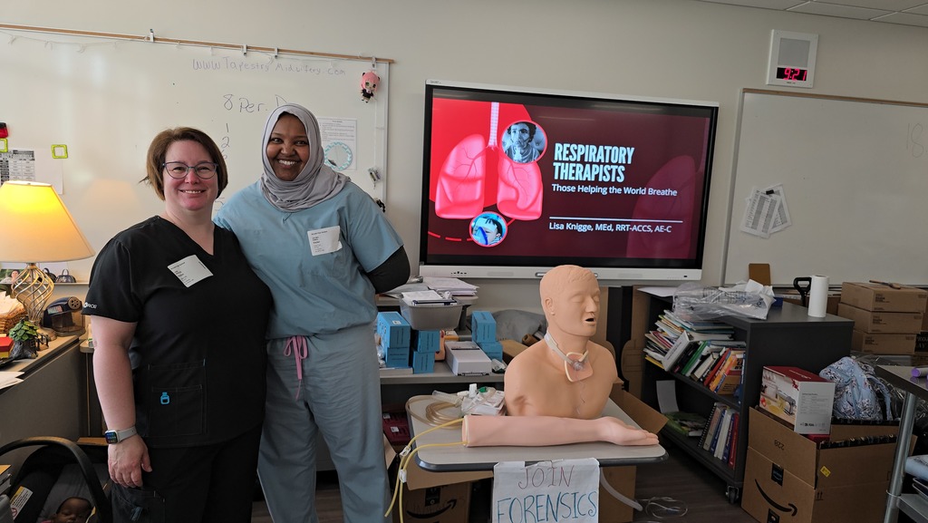Two women, one in scrubs and the other in a black top, standing next to a table with a mannequin.