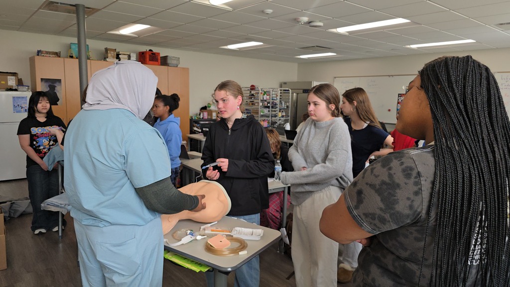 A group of people in a classroom, a person in blue scrubs demonstrates with a mannequin head.
