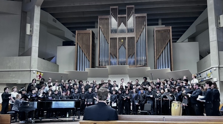 Choir performs in a large, well-lit room with a grand pipe organ at the back. Audience seated on benches.