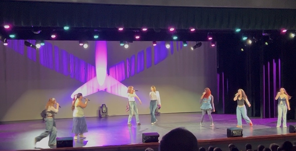 Several people perform on a stage with colorful lights in the background. They wear various colored pants and sneakers.