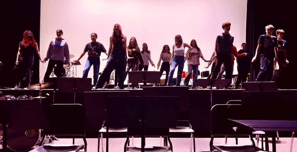 A group of people on a stage, performing in front of a large screen, with musical instruments and chairs behind them.