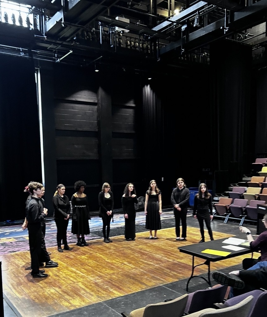 A group of individuals in black attire stands on a wooden stage, facing forward. Behind them, a row of chairs is arranged, and a desk with papers sits on the right.