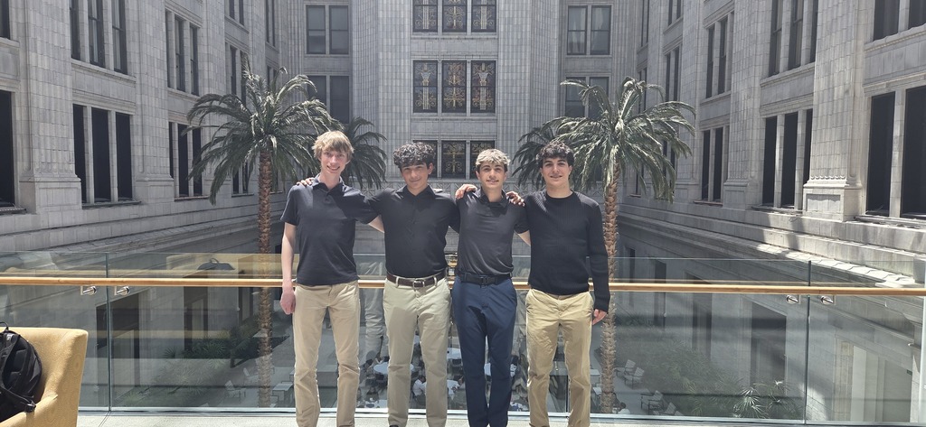 Four men stand together on a balcony in front of a building, wearing black shirts and pants. Behind them are palm trees.