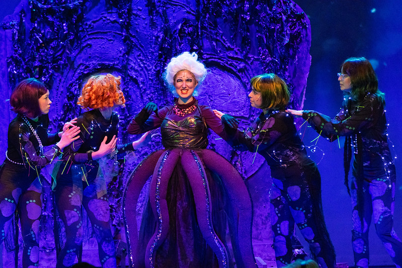 Five people in costumes stand on a stage with a purple background. One person wears a white wig.