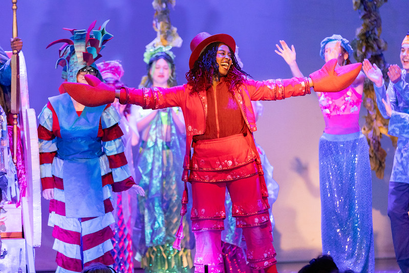 A performer in a red costume with arms extended on stage, surrounded by others in colorful costumes.