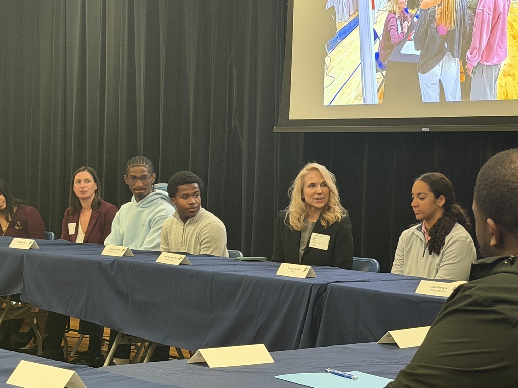 A group of individuals seated around a table in a room with a projector screen behind them.