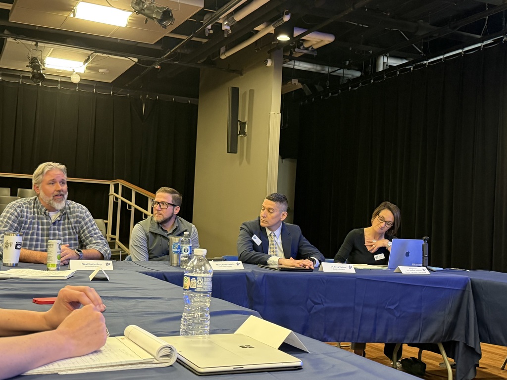 Five people are seated at a table, likely in a conference room, with laptops, a water bottle, and papers.