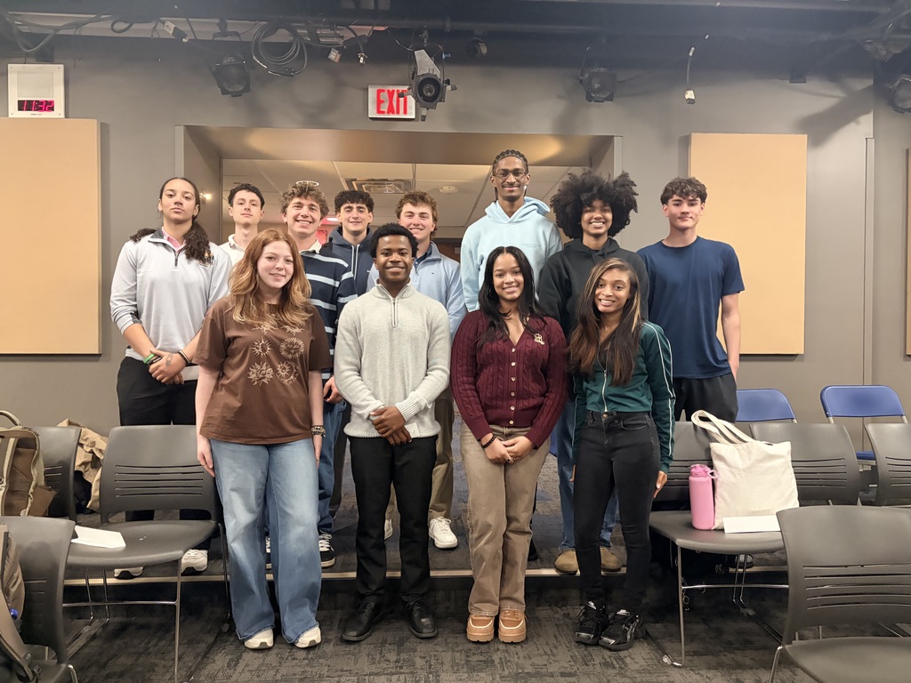 A group of young adults standing in a classroom with chairs and a table. An exit sign is visible above.