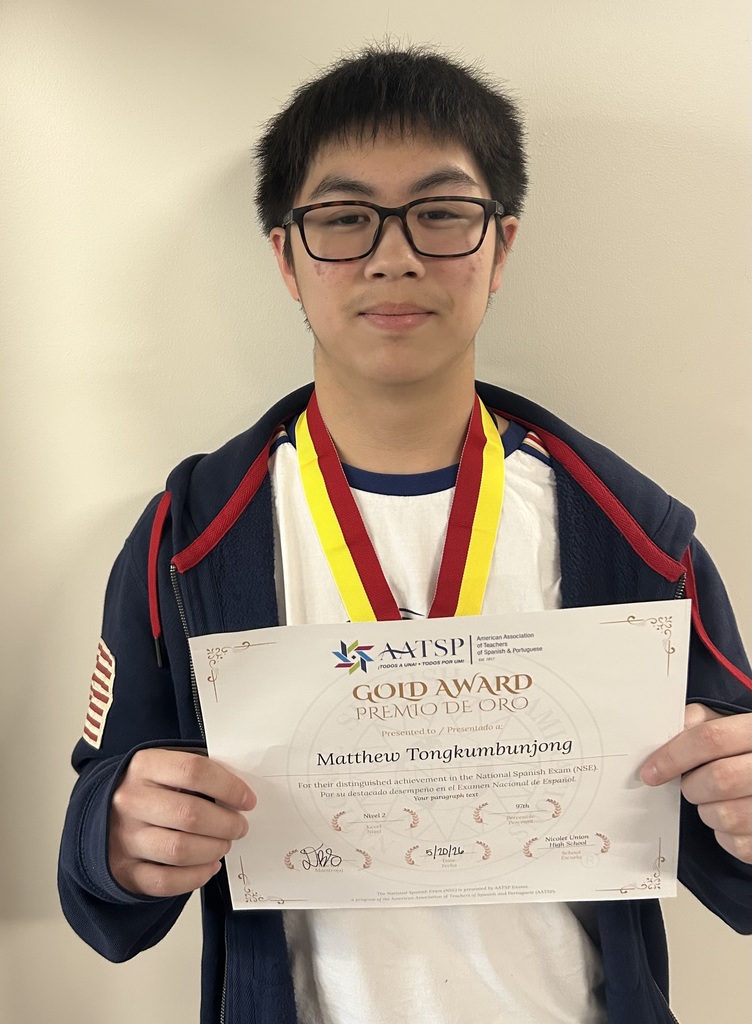 Person holding a paper with "GOLD AWARD" on it, wearing glasses and a jacket with a gold medal around the neck.