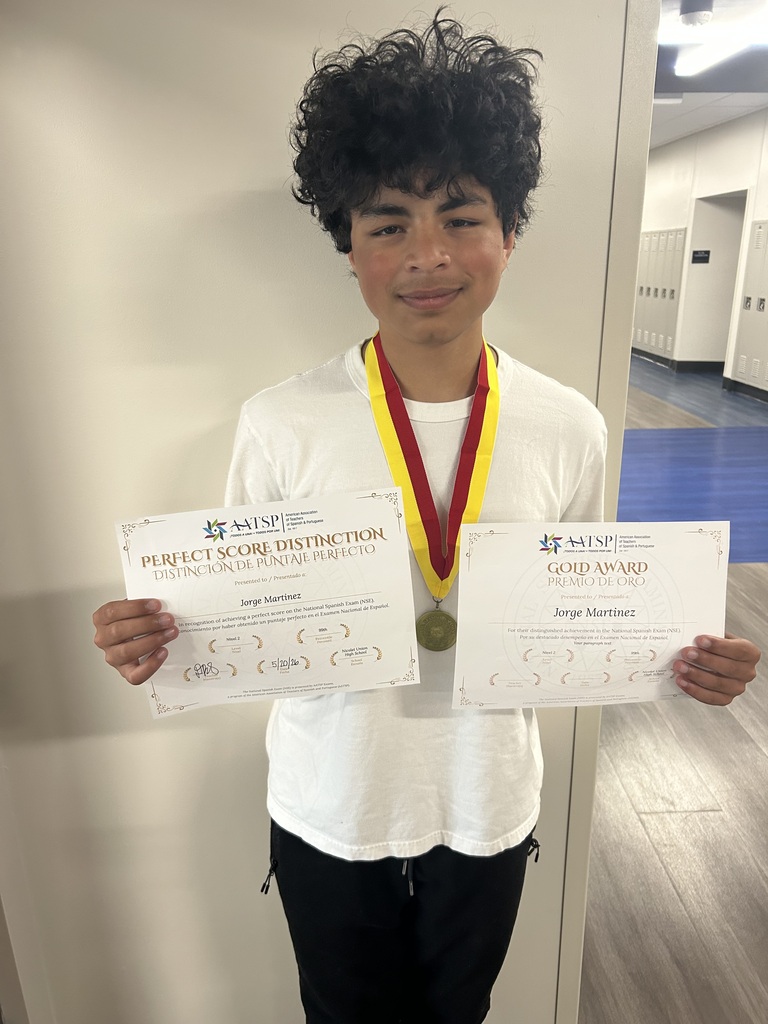 A young person displays two certificates and a medal in a corridor, wearing a white shirt and black pants.