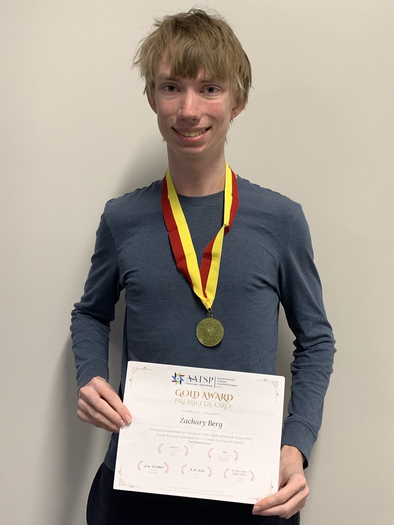 A young person in a gray shirt holds a certificate and a gold medal, smiling at the camera.