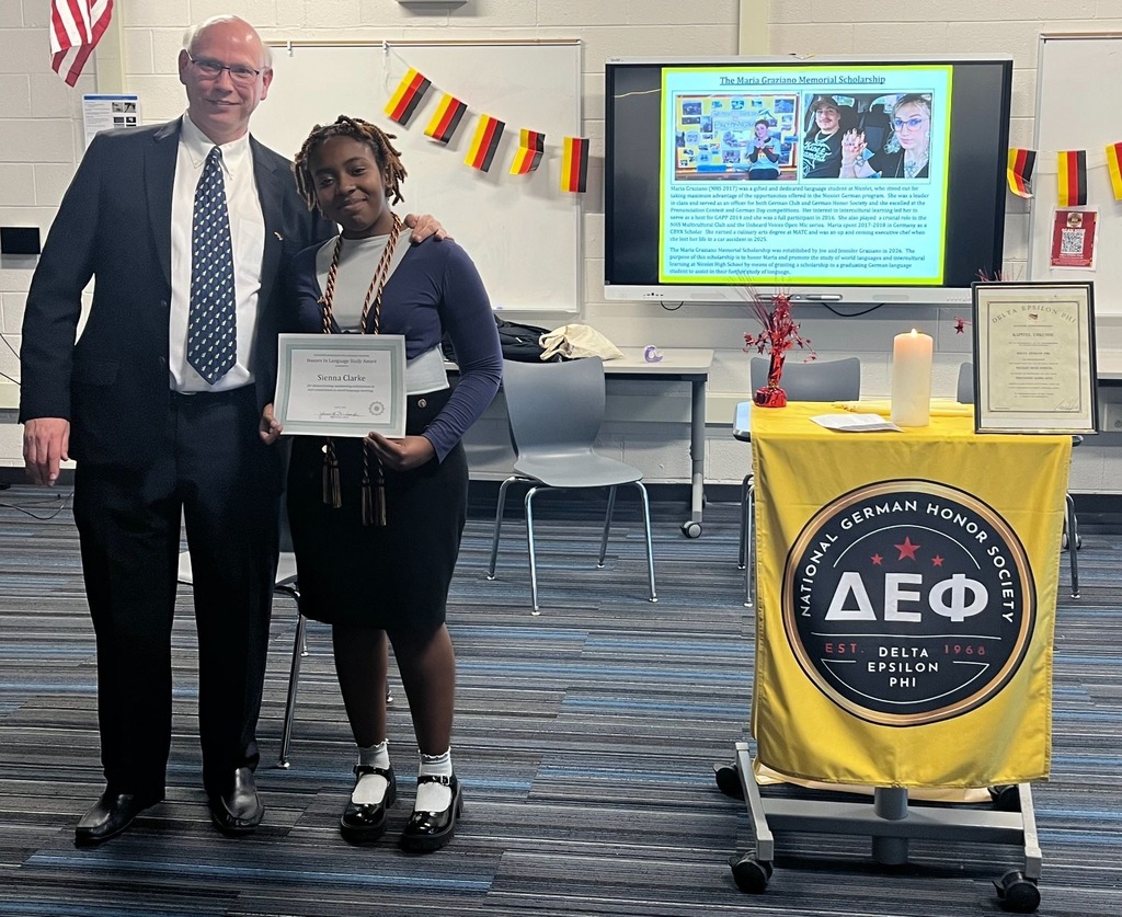 A man and a woman stand next to each other holding a framed award. They are in a classroom with a presentation screen and a desk.