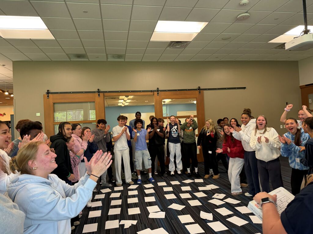 A room with several people, some standing and others sitting, clapping. A person holds papers on a table.