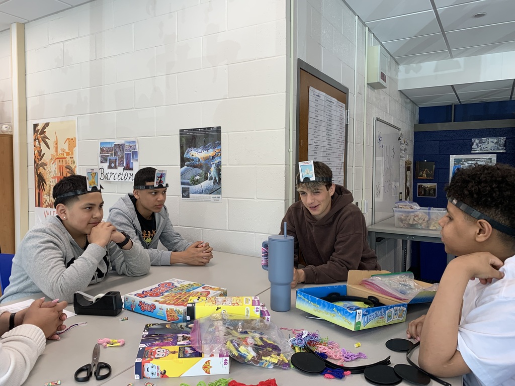 Four people sit around a table with craft supplies. Two wear caps, one wears glasses, and another wears a watch.